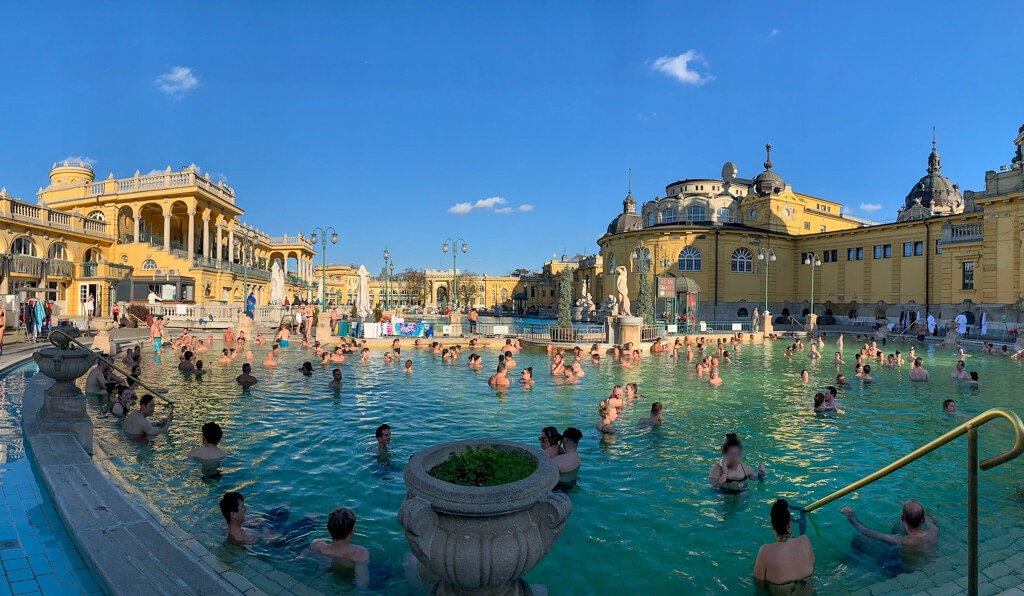 View of the open air thermal baths in Budapest with many people enjoying the warm water under a clear blue sky.