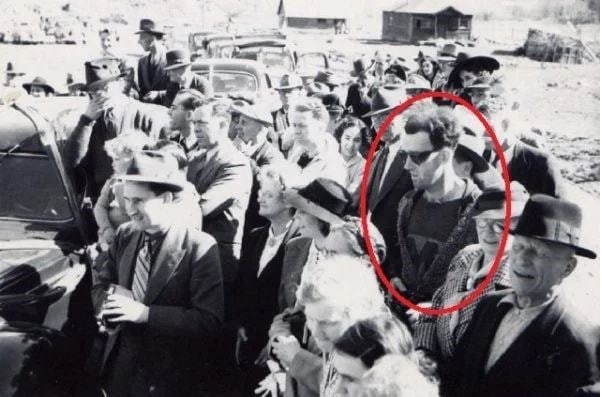 A black and white historical photo featuring a crowd of people, with one individual dressed in distinctly modern clothing amidst the period attire, fueling time traveler speculation.