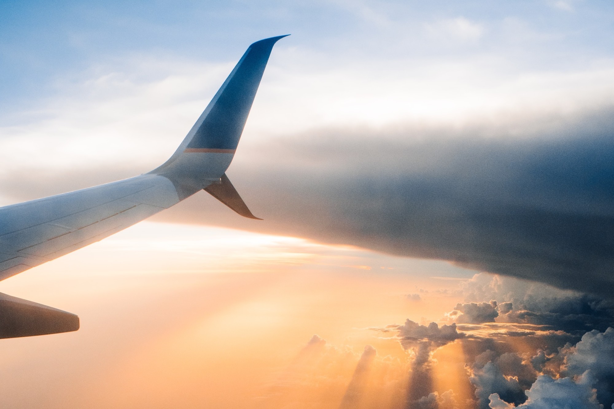 The wing of a passenger jet is visible from the view of an airline seat. Brilliant rays of sunshine also break through dark clouds