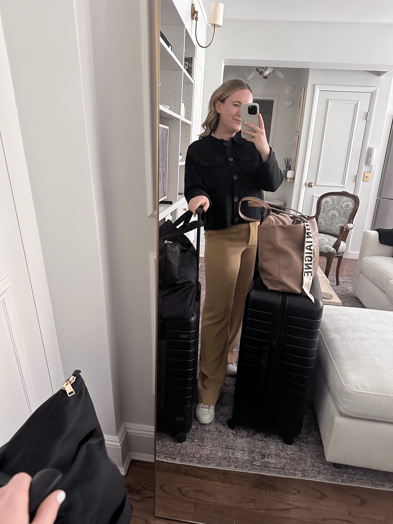 Woman wearing a casual travel outfit consisting of a beige sweater and black trousers, standing in front of a blurred outdoor background, illustrating a simple yet stylish look for airplane travel