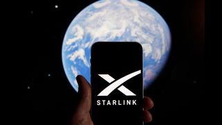 In the foreground is a hand holding a phone with the Starlink logo on the screen behind it in the background of the image is planet Earth.