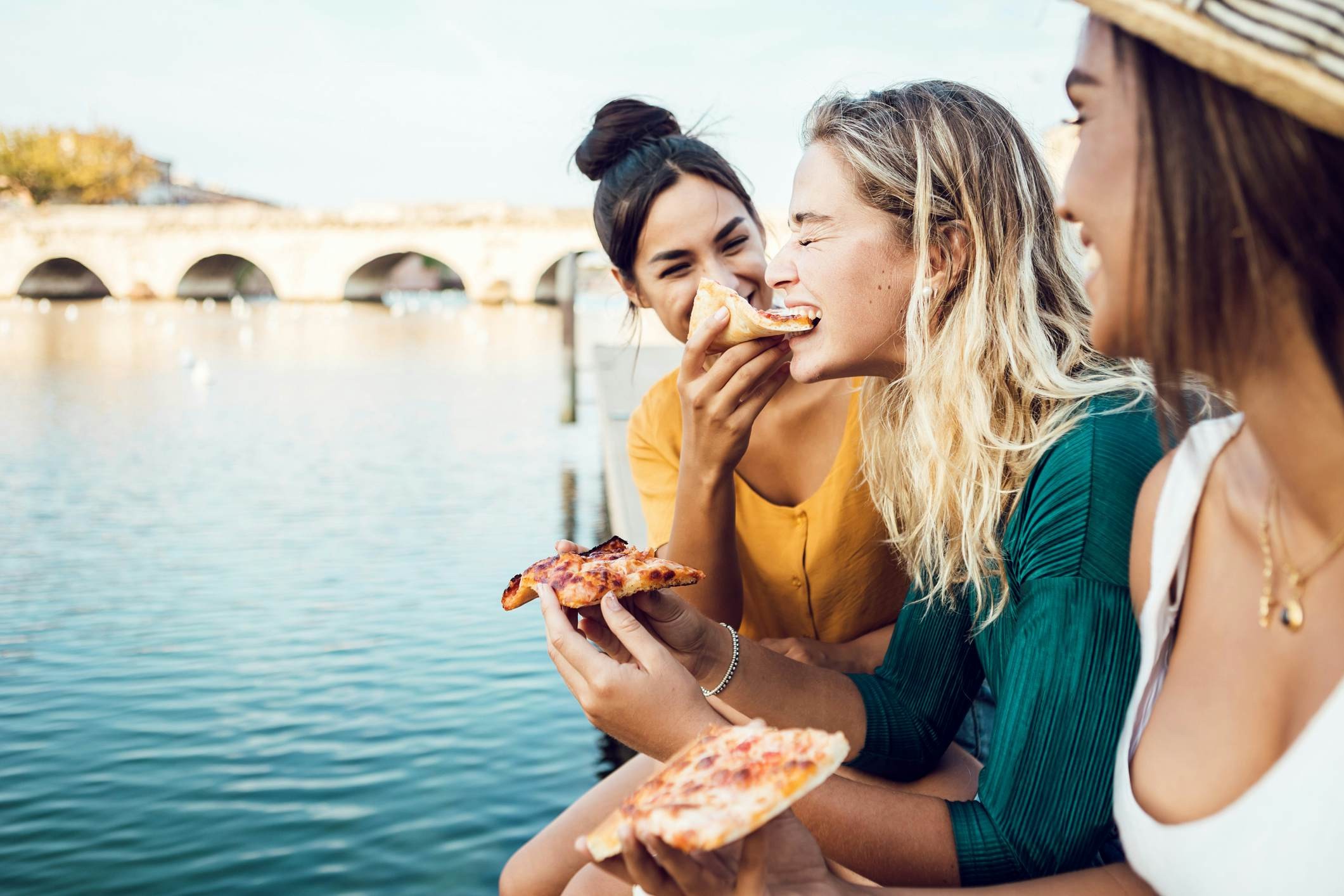 Friends sharing pizza by a river in Italy, highlighting affordable food options
