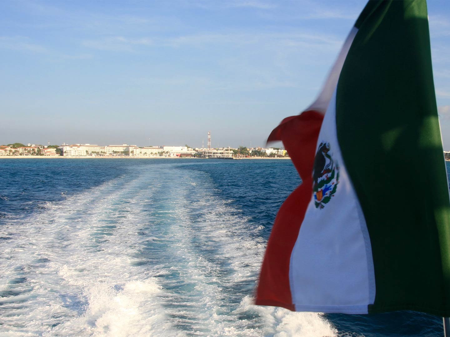 A Mexican flag flutters in the wind looking towards the coastline as a boat sails off into the sea.