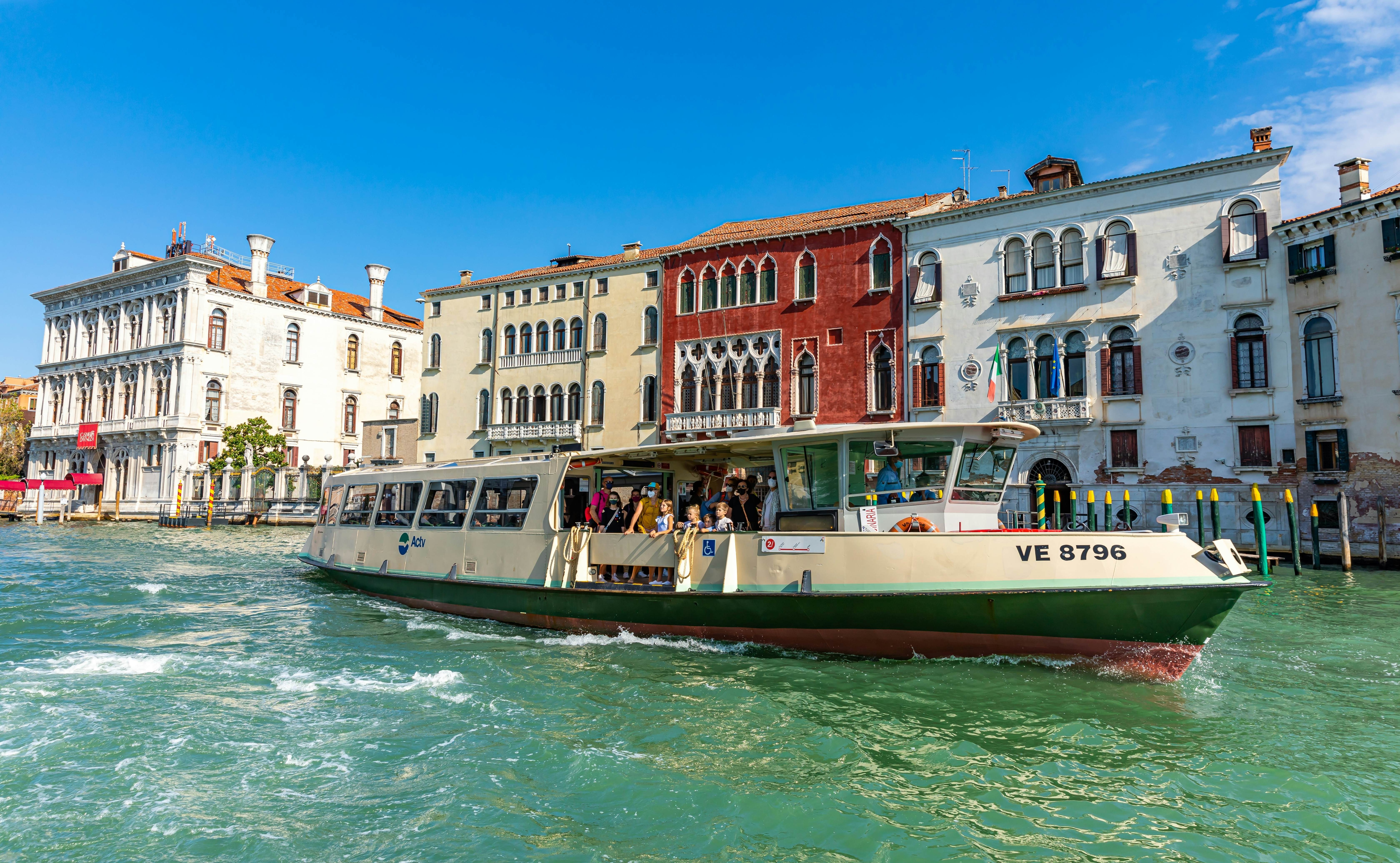 A vaporetto public ferry sets off in a green-tinted canal, with colorful palaces with Gothic arches seen in the background