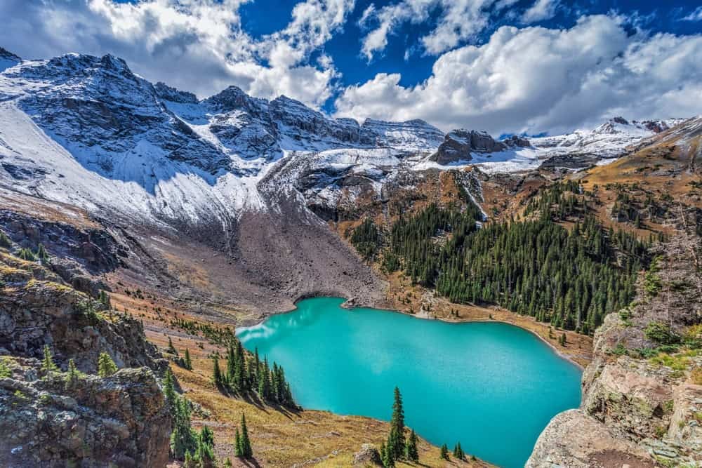 A scenic view of Blue Lakes shows a turquoise lake surrounded by mountains and forests under a blue sky.