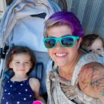 Woman in sunglasses smiling with baby on back in carrier and toddler in uppababy minu stroller in a bus stop