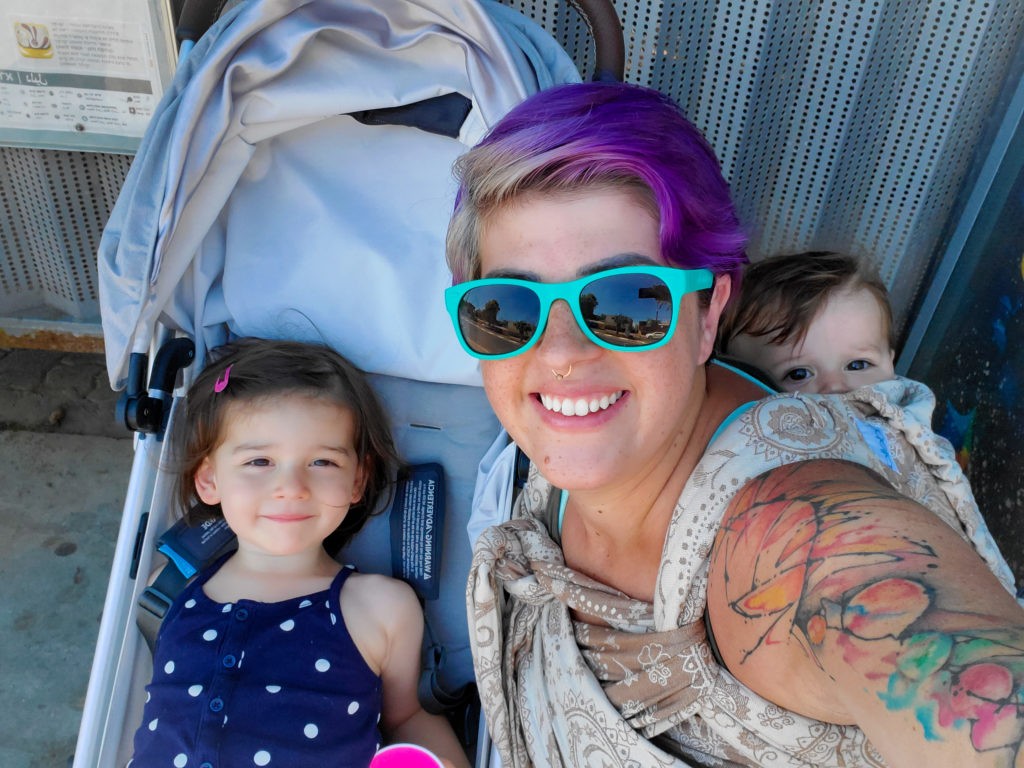Woman in sunglasses smiling with baby on back in carrier and toddler in uppababy minu stroller in a bus stop