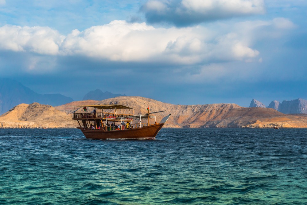 Dhow boat Cruise in Arabian Peninsula, Oman. A boat on blue water with desert mountains in the background.