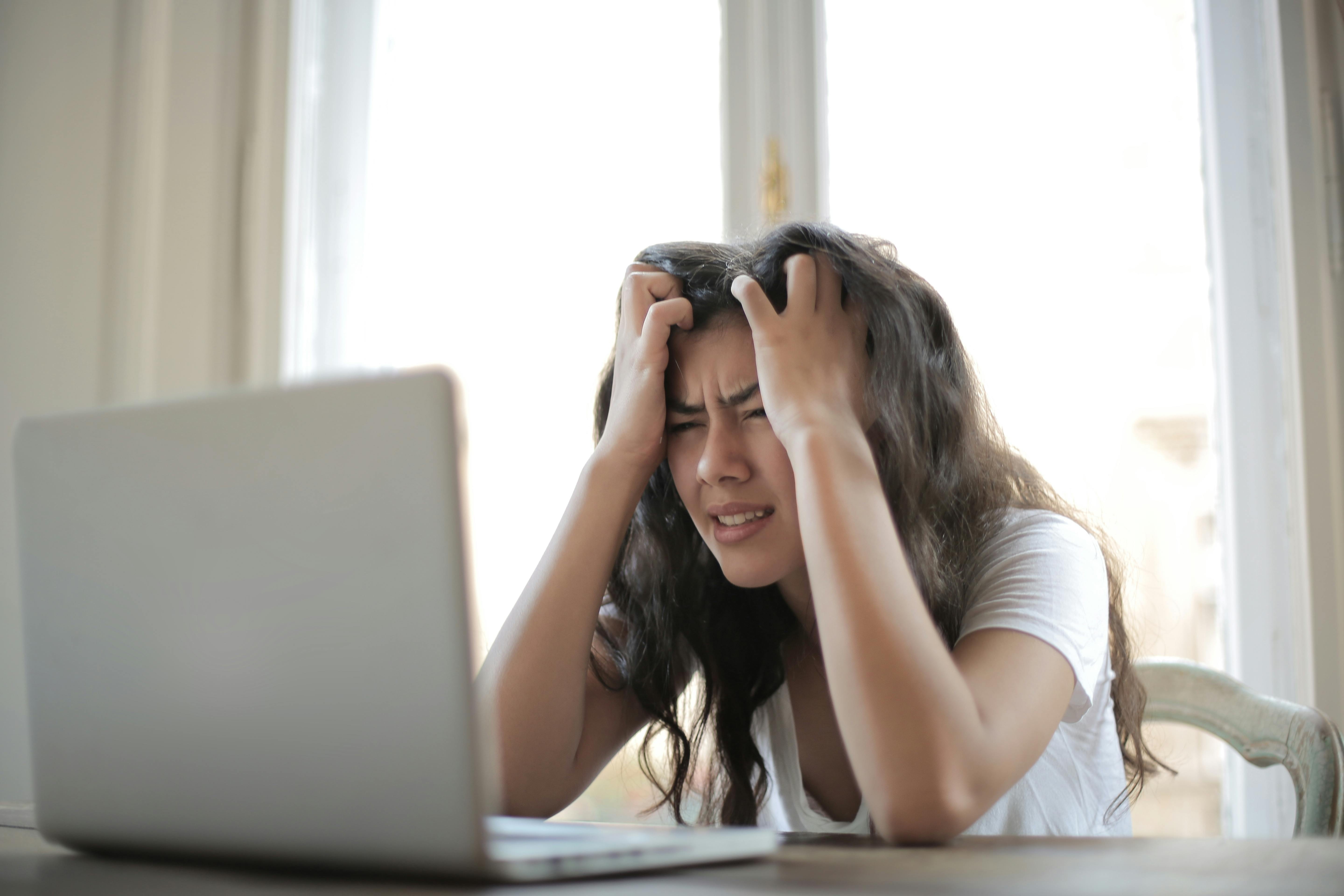A frustrated woman with long curly hair holds her head in her hands while looking at a laptop screen in a bright room.