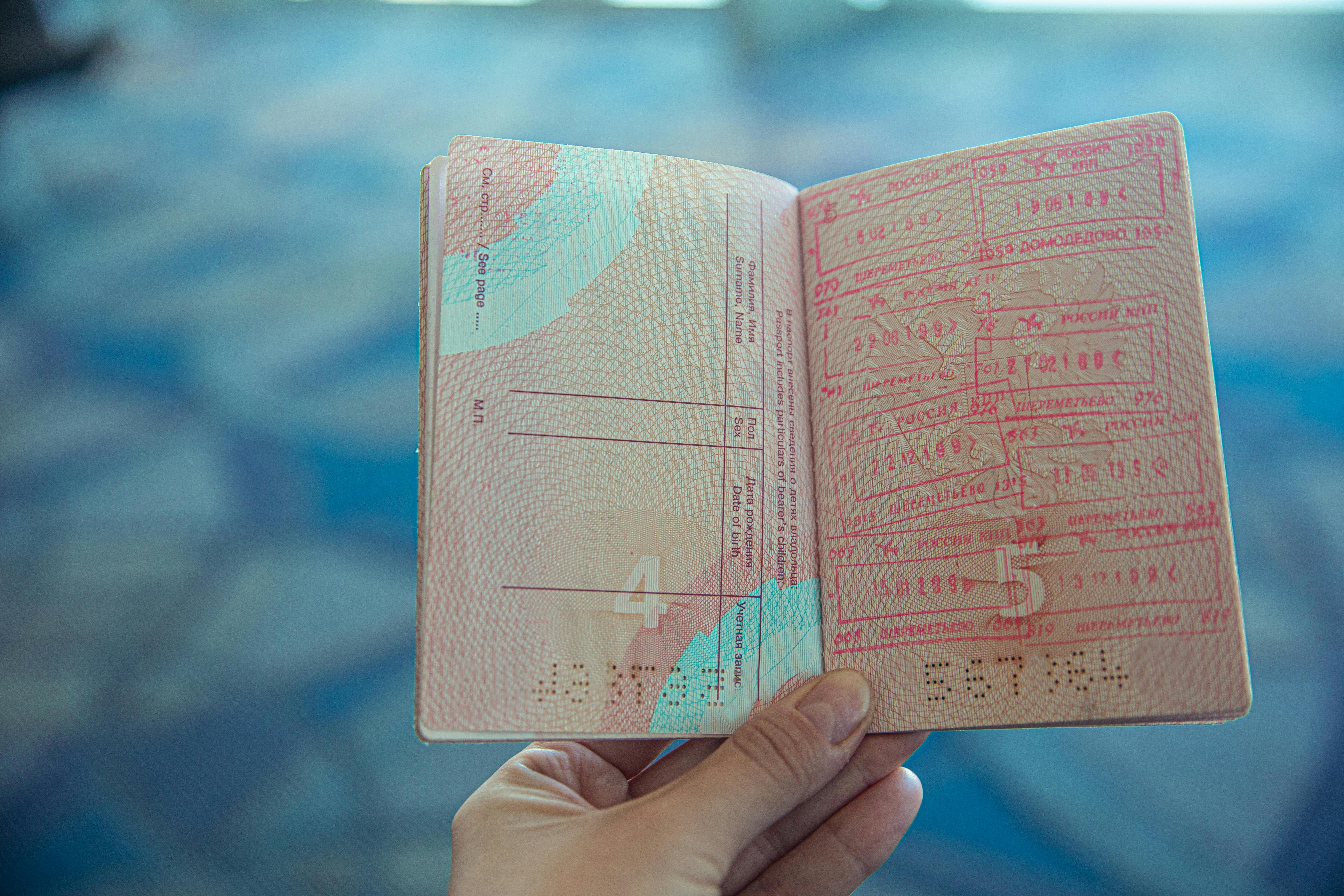 A person holding an open passport with visa stamps and entry-exit markings, against a blurred blue-toned background.